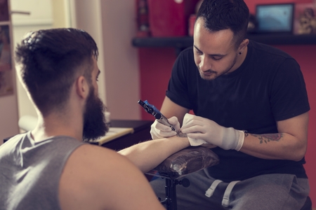 Male tattoo artist holding a tattoo gun, showing a process of making tattoos on a male tattooed model's arm.の写真素材