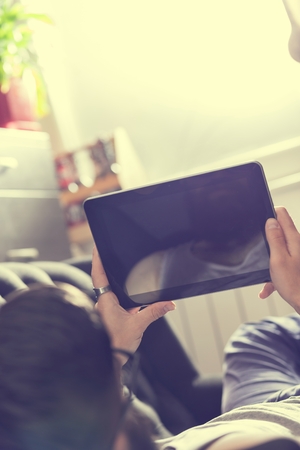 Male model lying on a couch in a living room, surfing the web on a tablet computerの写真素材