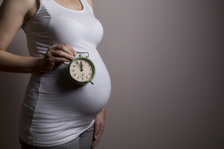 Close up of a pregnant woman's belly, woman standing next to a grey background and holding a clock representing a proximity of a due dateの写真素材