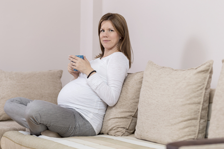 Pregnant woman sitting on a couch in a living room, holding a cup of teaの写真素材