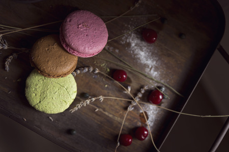 Top view of macaron cookies on a wooden tray, with cherry fruit and lavender flowers placed next to it. Selective focus on brown and pink cookieの写真素材