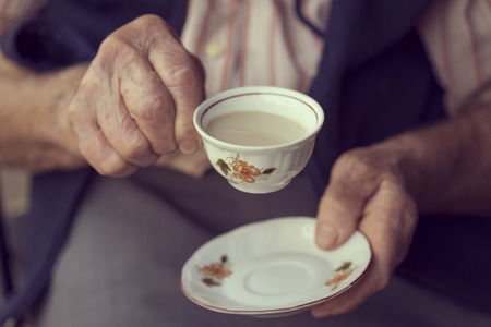 Detail of an elderly woman's hands, holding a rustic cup filled with coffee with creamの写真素材
