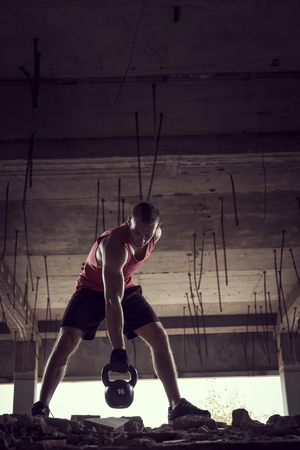 Front view of a young muscular athlete working out, lifting a kettlebell weight in an abandoned ruined buildingの写真素材