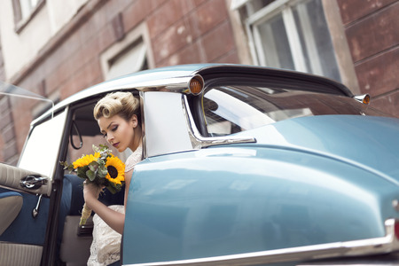 Beautiful young bride sitting in a wedding dress in a retro old car, holding a sunflower bouquetの写真素材