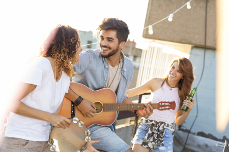 Three young friends having fun at rooftop party, playing guitar and singing. Focus on the man playing the guitarの写真素材
