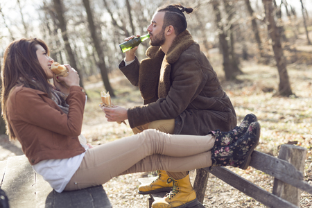 Young couple sitting on a wooden bench by the forest road, taking a break while eating sandwiches and drinking beer. Focus on the guyの写真素材