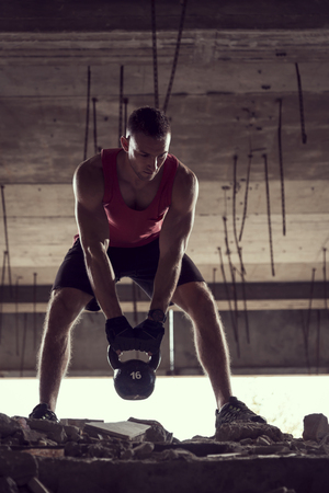 Front view of a young muscular athlete working out, lifting a kettlebell weight in an abandoned ruined buildingの写真素材