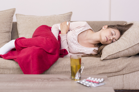 Young woman lying on a bed covered with blanket, holding her stomach in painの写真素材