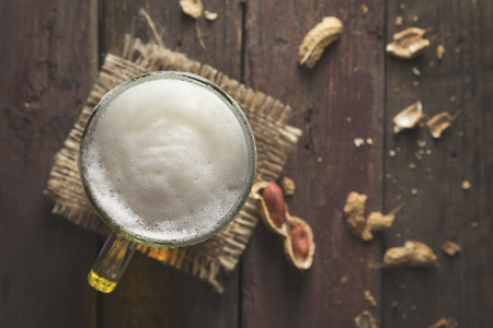 Top view of a mug of pale beer placed on a burlap cover with froth and some peanuts on a rustic wooden pub table. Focus on the frothの写真素材