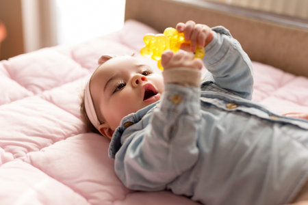 Adorable baby girl lying on a pink duvet on bed, holding a rattle and playing