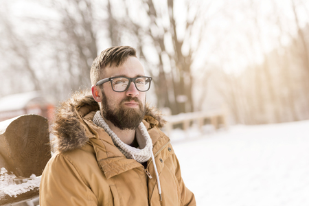 Handsome young man wearing a warm winter clothes, leaning against a wooden fence and enjoying a snowy winter dayの写真素材