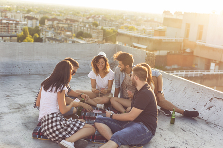 Group of young people sitting on a picnic blanket, having fun while playing cards on the rooftop. Focus on the couple in the middleの写真素材