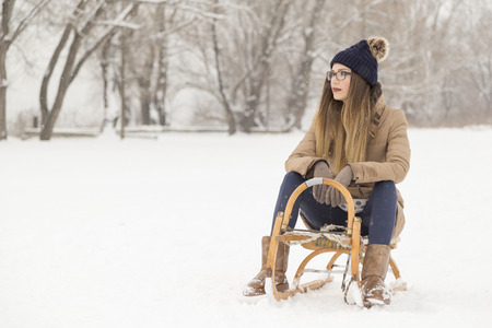 Beautiful young woman wearing a warm winter clothes and sitting on a sleight on a snowy winter dayの写真素材