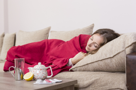 Sick woman covered with a blanket lying in bed with high fever and a flu. Pills, teapot and lemon on the tableの写真素材