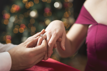 Young attractive couple in love sitting at a dinner table next to a nicely decorated Christmas tree while man surprises his girlfriend with an engagement ringの写真素材