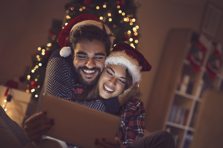 Couple in love sitting next to a Christmas tree, wearing Santa's hats and having fun looking at old photos on a tablet computerの写真素材