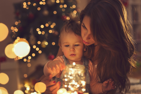Beautiful young mother and daughter sitting on the floor next to a Christmas tree, hugging and holding a jar of Christmas lights.の写真素材