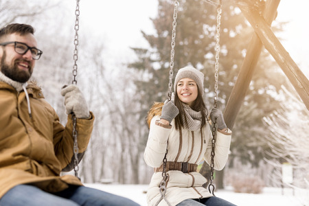 Beautiful young couple sitting on a swing and having fun on a snowy winter dayの写真素材
