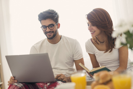 Beautiful young couple in love having fun reading and surfing the net while having breakfast in bed. Focus on the guyの写真素材