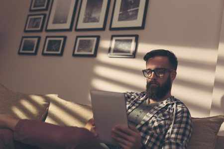 Handsome young man sitting on a living room sofa, working on a tablet computerの写真素材