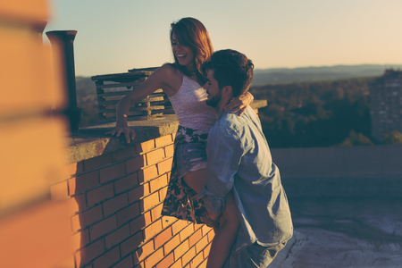 Couple in love having fun on a building rooftop, enjoying a beautiful summer sunset over the city; guy lifting the girlの写真素材