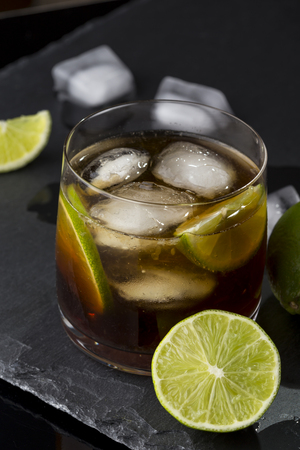 High angle view of a glass of Cuba Libre cocktail with rum, coke, lemon juice and ice cubes on modern black stone tray. Selective focusの写真素材