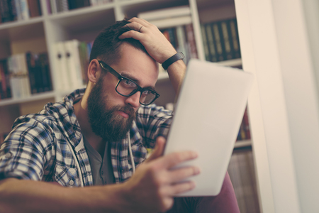 Handsome man sitting on the floor of his home library, surfing the net on a tablet computer, relaxing on his leisure timeの写真素材