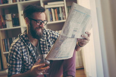 Handsome young man reading newspapers and having a drink in his home libraryの写真素材