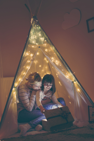 Mother and daughter sitting in a teepee enlightened by Christmas lights, opening a present. Focus on the motherの写真素材