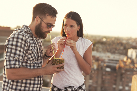 Young couple in love drinking a pineapple cocktail and having fun at rooftop party in sunset. Focus on the guyの写真素材
