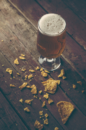 Glass of cold pale beer with some tortilla chips on a rustic wooden table. Selective focus on the foamの写真素材