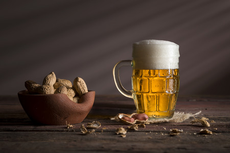 Mug of light beer with foam placed on a burlap coaster and a bowl of peanuts on a rustic wooden tableの写真素材