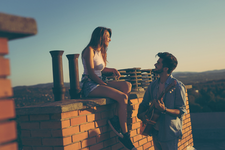 Guy playing the guitar to his on a building rooftop; couple at a summer rooftop party enjoying sunset over the cityの写真素材