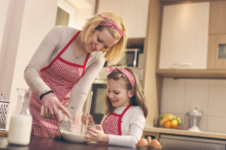 Mother and daughter baking dough in the kitchen; mother adding flour from a jar while daughter stiring with a kitchen spoon. Focus on the daughterの写真素材