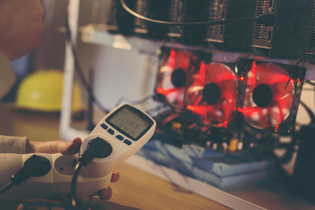Detail of male hand holding a wattmeter measuring cryptocurrency mining rig electrical energy consumption. Selective focus on the wattmeterの写真素材