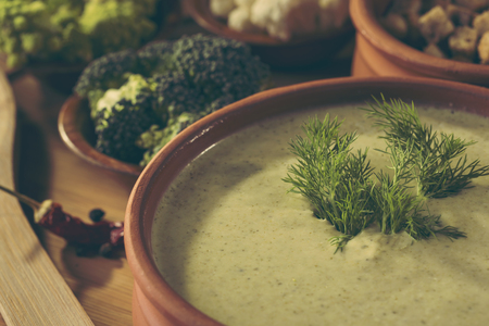 High angle view of fresh creamy broccoli soup with some fresh vegetables, spices and croutons next to it. Selective focus on the dill leavesの写真素材