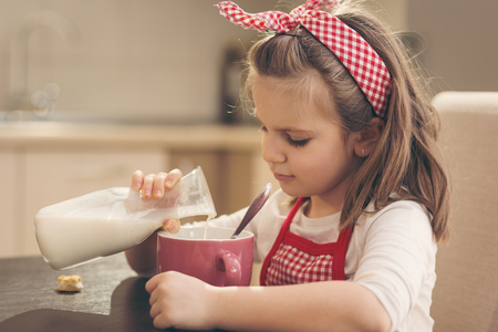 Little girl sitting in the kitchen, having breakfast, pouring milk into a cereal cup, preparing herself a healthy, tasty mealの写真素材
