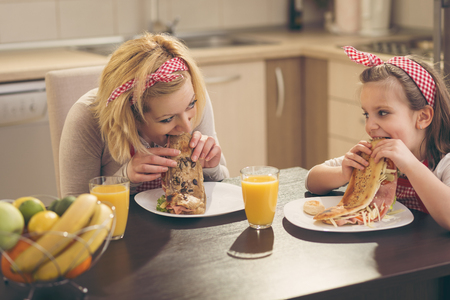 Beautiful mother and daughter in the kitchen having breakfast, eating sandwiches and drinking orange juice. Focus on the motherの写真素材