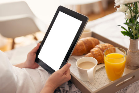 Detail of female's hands holding a black tablet computer with blank white screen, sitting in bed and having breakfast. Selective focus on the tabletの写真素材