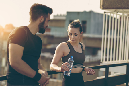Young couple standing on a building rooftop terrace, drinking water and speaking while taking a break from a workoutの写真素材