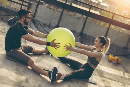 Couple sitting on a yoga mat on a building rooftop terrace, exercising with a pilates ballの写真素材