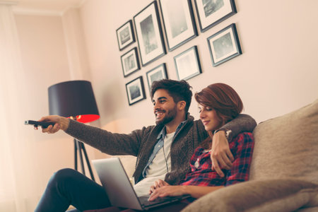 Couple in love sitting on a living room sofa, watching TV. Man using a remote control while woman is...