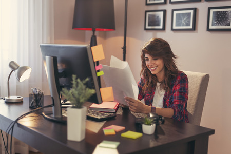 Young woman sitting at a desk at her home office, holding documents joyful after a business successの写真素材