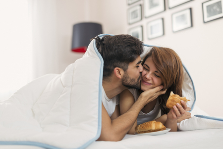 Couple in love lying under the sheets, having breakfast in bed and kissingの写真素材