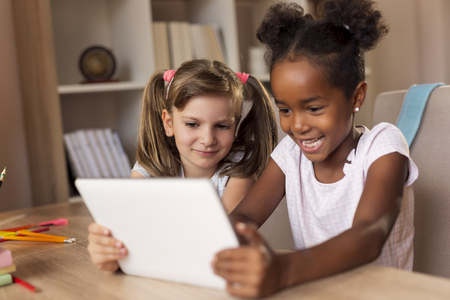 Two little girls sitting at a desk, playing video games on a tablet computer and having fun. Focus on the girl on the leftの写真素材