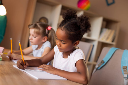 Two little girls sitting at a desk, writing in their notebooks, doing a math homework and studying...