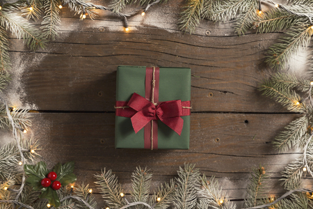 Top view of a frame made of snowy pine branches and Christmas lights with a nicely wrapped gift box placed on wooden tableの写真素材