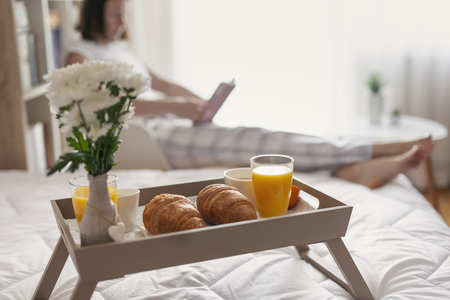 Woman wearing pajamas, sitting in a chair next to bed, reading a book. Breakfast tray in the foreground. Selective focus on the right croissantの写真素材