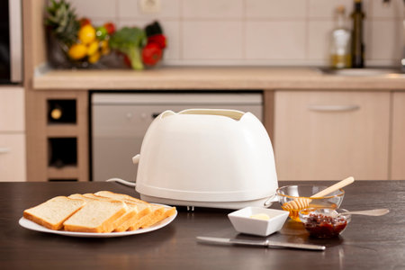 Toaster on the kitchen counter, with toasted bread and bowls with butter, jam and honey. Focus on...
