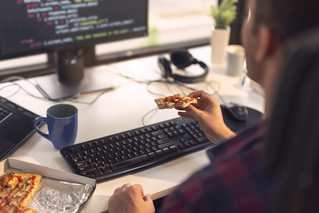 Software developer sitting at a desk in a software developing office, eating pizza and drinking coffee on a lunch break. Focus on the pizzaの写真素材